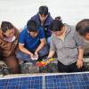 Several people look at an electrical device that’s connected to a solar panel.